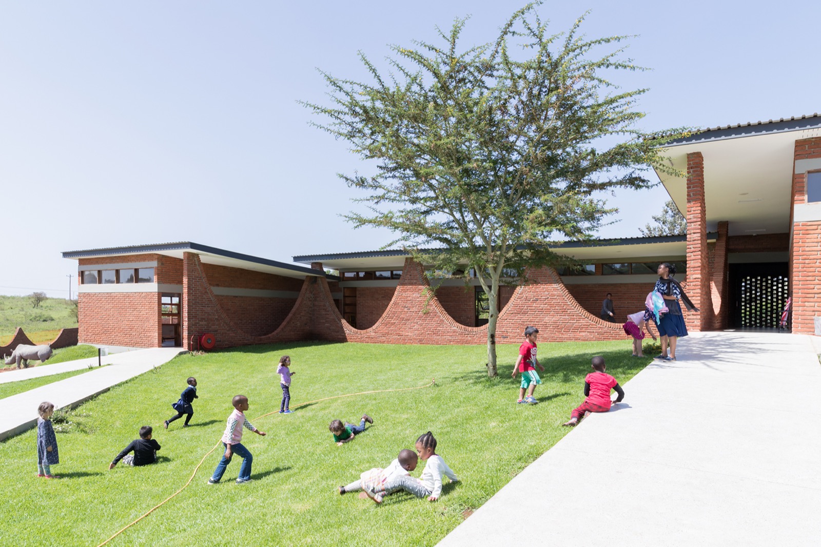 Backstein-Klassenzimmer der Black Rhino Academy in Karatu, Tansania, mit einer geschwungenen Kettenbogen-Wand und auf dem Rasen spielenden Kindern