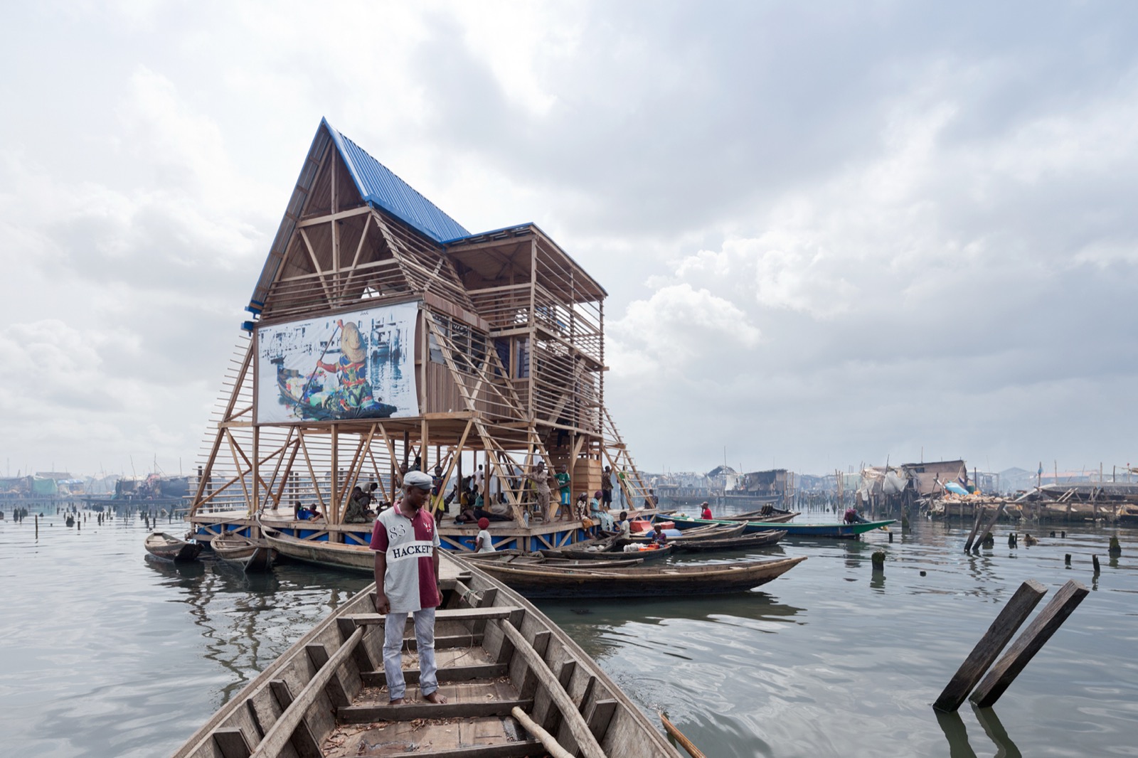 Die Makoko Floating School, eine A-Rahmen-Konstruktion aus Holz, die auf Plastikfässern auf der Lagune von Lagos schwimmt, mit Kanus und der Pfahlbausiedlung dahinter