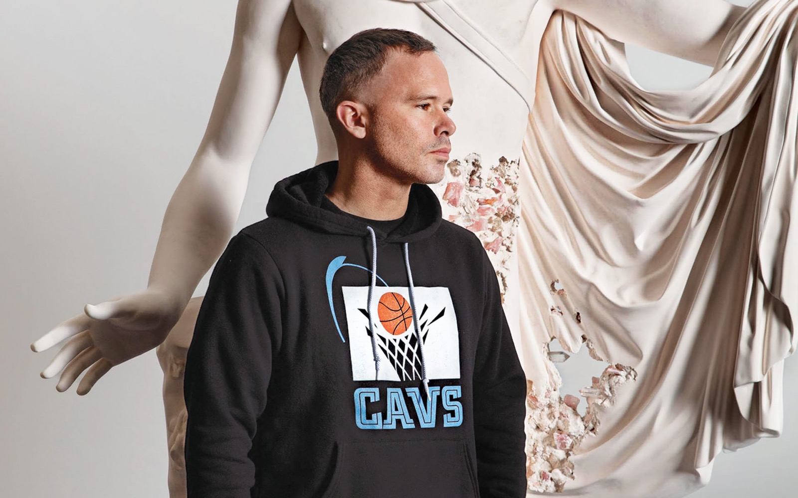 Daniel Arsham com um moletom do Cleveland Cavaliers ao lado de uma reprodução erodida de uma escultura clássica drapeada