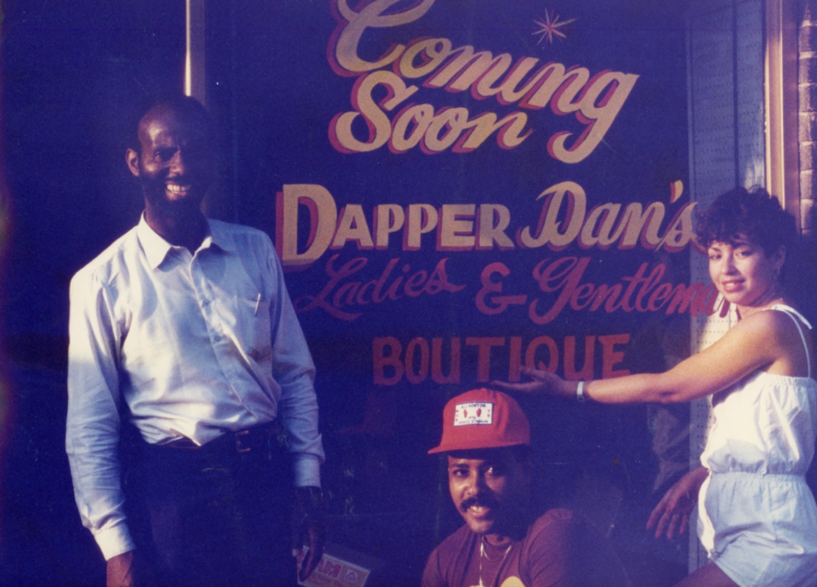 A vitrine pintada à mão da Dapper Dan's Boutique no Harlem, com os dizeres "Dapper Dan's Ladies & Gentlemen Boutique", com pessoas posando do lado de fora