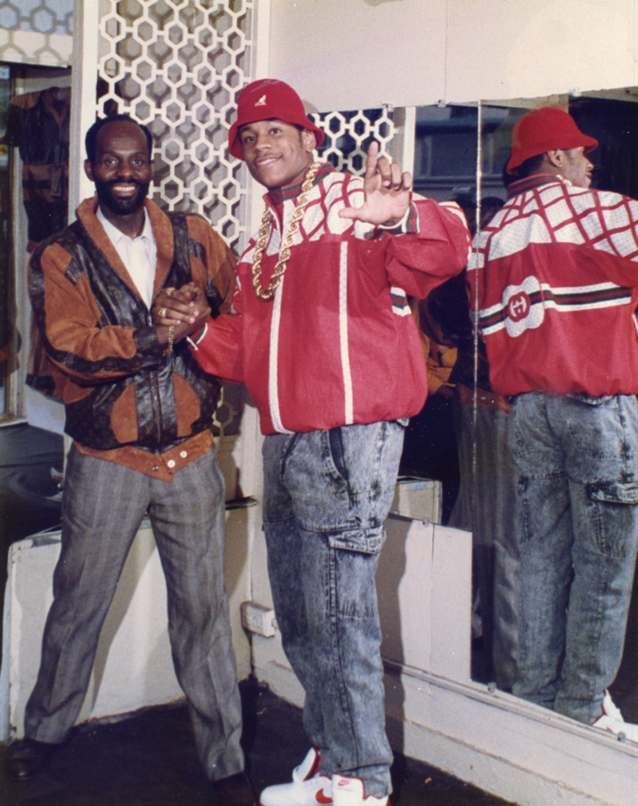LL Cool J vestindo um agasalho vermelho com listras da Gucci, o logo da Gucci visível no espelho atrás dele, ao lado de Dapper Dan em frente à boutique do Harlem