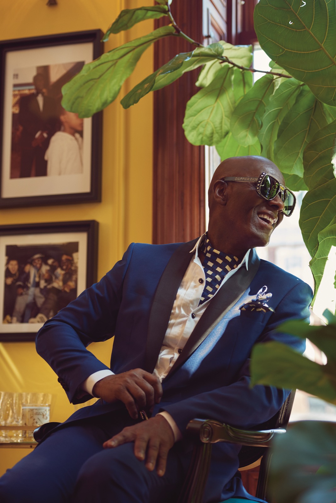Dapper Dan com uma jaqueta de smoking azul e óculos escuros ornamentados, sentado em seu ateliê no Harlem com fotografias emolduradas de seu trabalho atrás dele