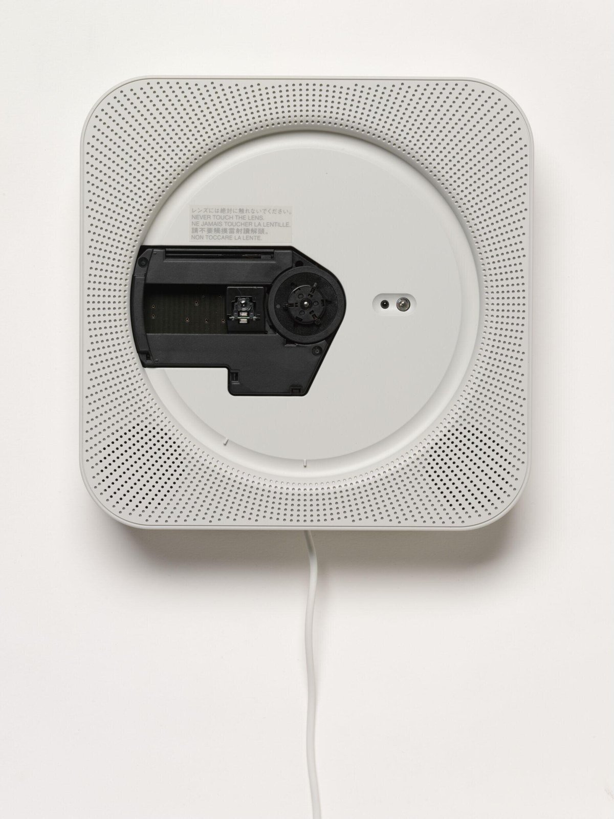 MUJI's wall-mounted CD player designed by Naoto Fukasawa in 1999, photographed straight-on for the V&A collection: a white square unit with perforated speaker holes radiating around the circular recess, the disc mechanism exposed, and the power cord hanging straight down as a pull switch