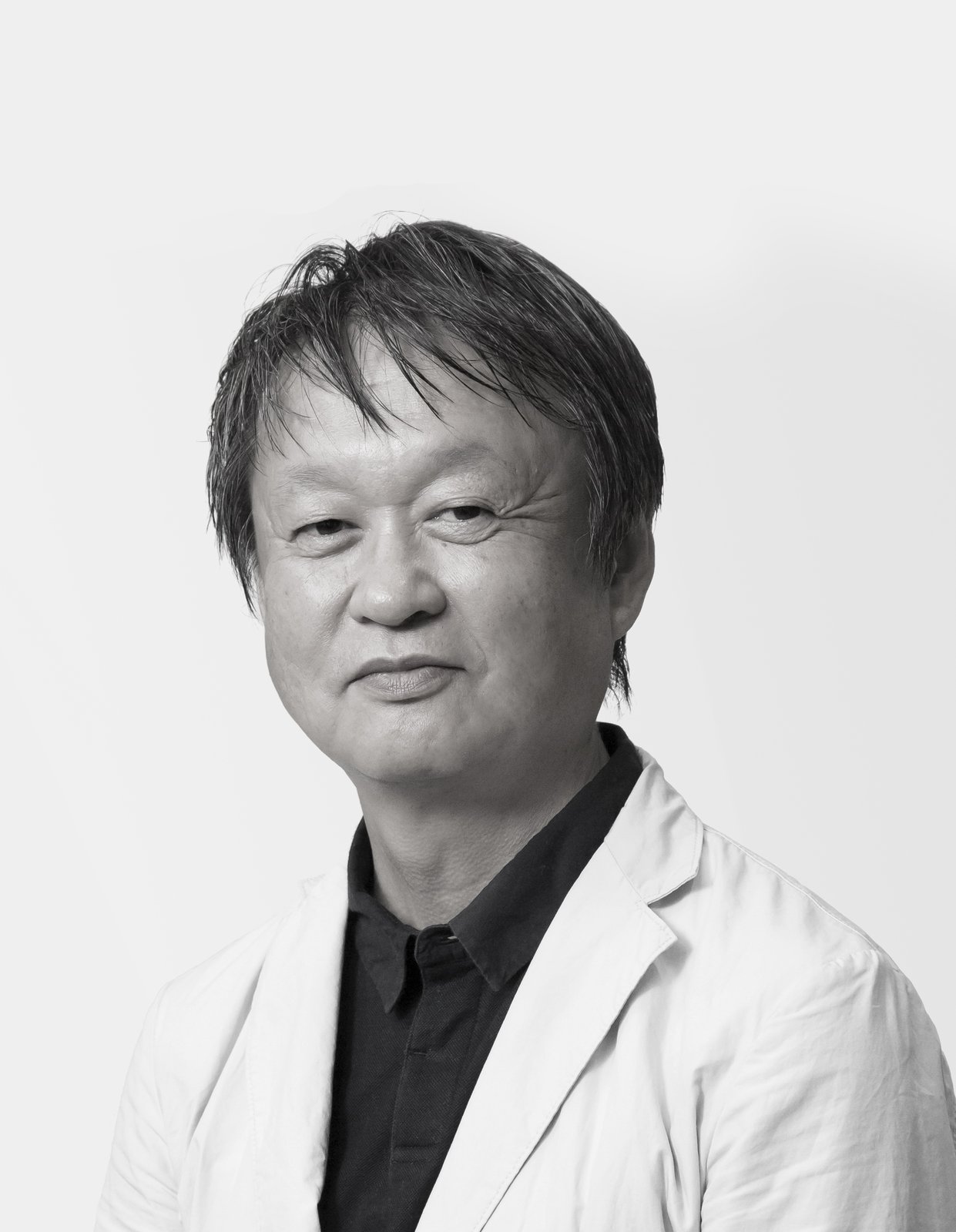Black-and-white studio portrait of Naoto Fukasawa from his official site: gray hair falling across his forehead, wearing a dark shirt under a pale jacket, against a plain white background