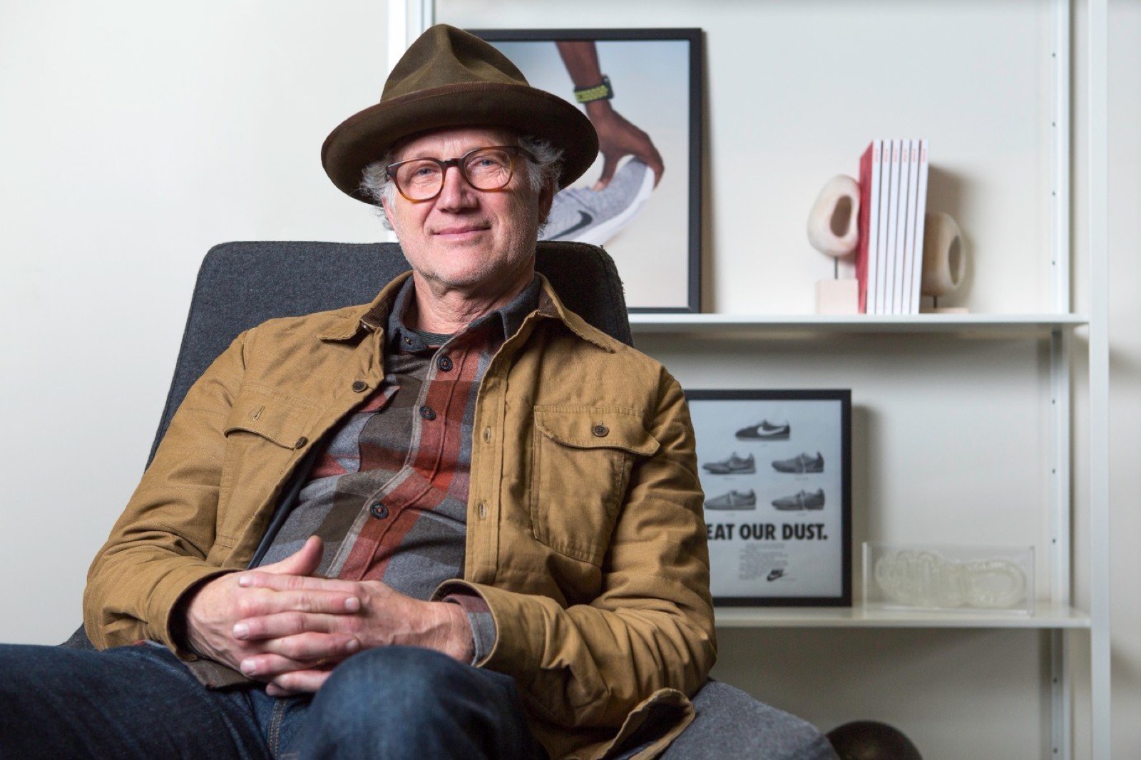 Tinker Hatfield sentado diante de designs de calçados Nike emoldurados e um anúncio vintage da Nike "Beat Our Dust"