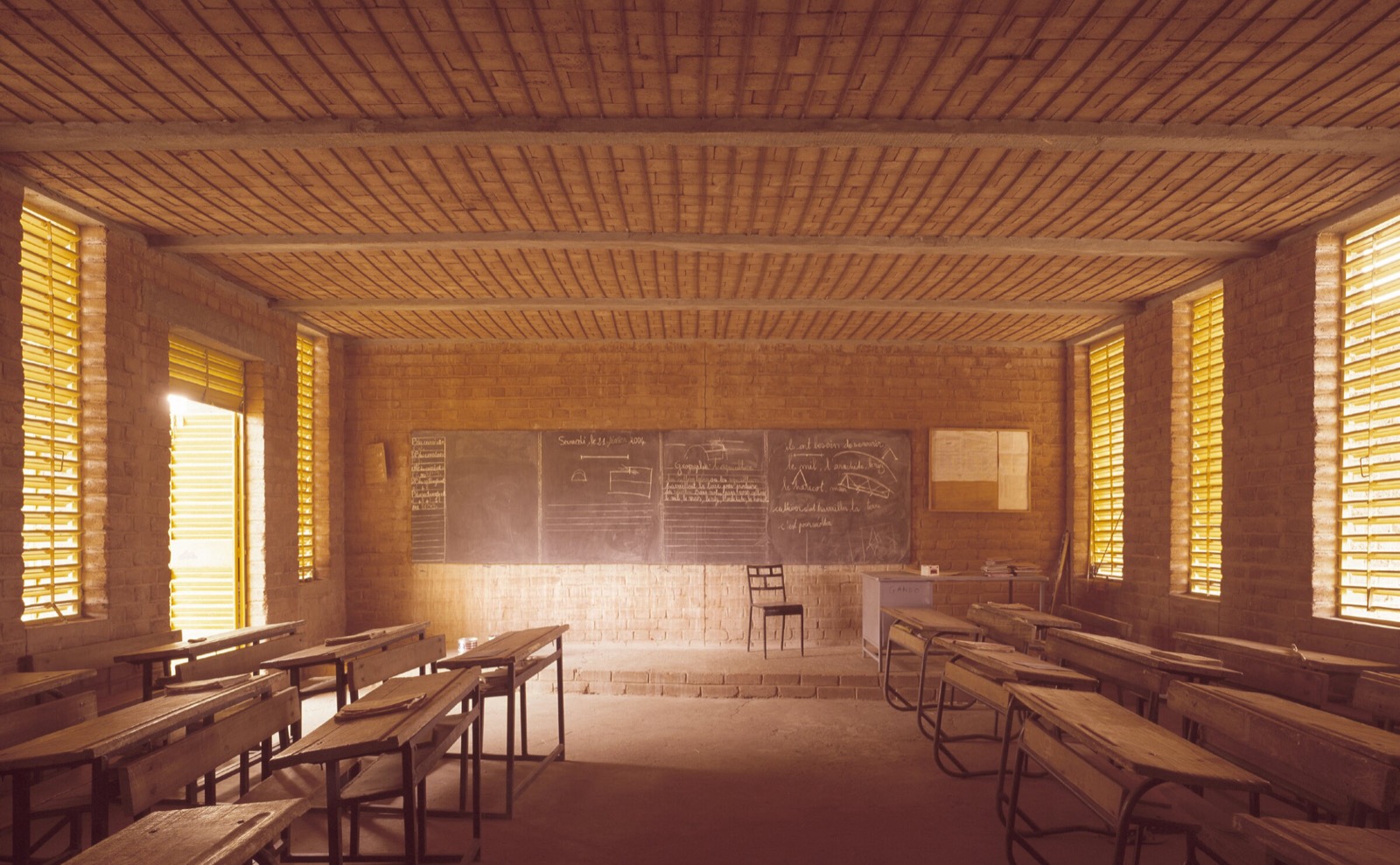 Innenraum eines Klassenzimmers der Gando Primary School, mit einer Decke aus Lehmziegeln, einer Tafel und perforierten Fensterläden, die Tageslicht auf die Tische filtern