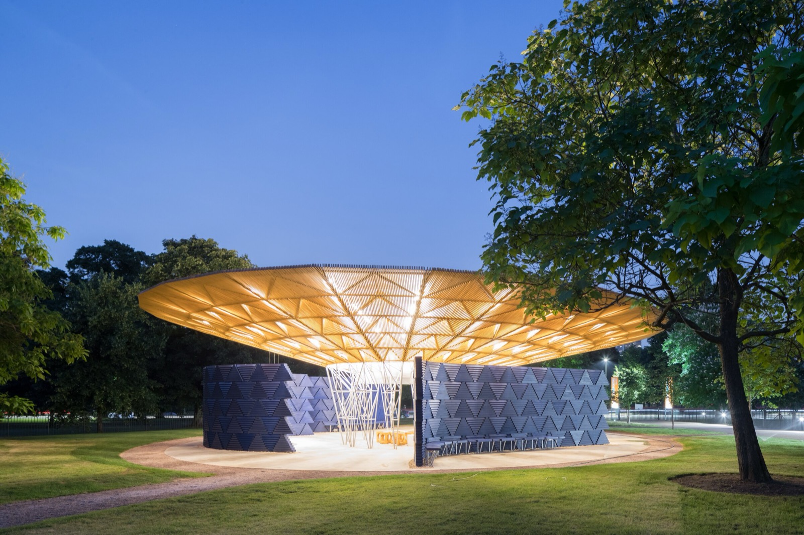 Serpentine Pavilion 2017 von Francis Kéré in den Kensington Gardens, London, mit seinem freistehenden Vordach, das in der Dämmerung über indigofarbenen modularen Wänden beleuchtet ist