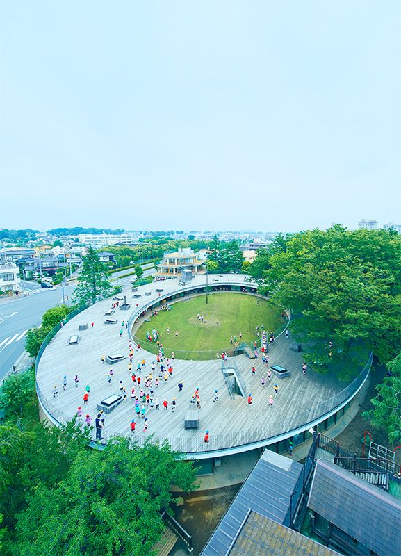 Vista aérea do Fuji Kindergarten em Tachikawa, Tóquio -- o prédio oval em forma de rosca da Tezuka Architects com crianças correndo em seu deck de telhado de madeira aberto ao redor de um pátio central