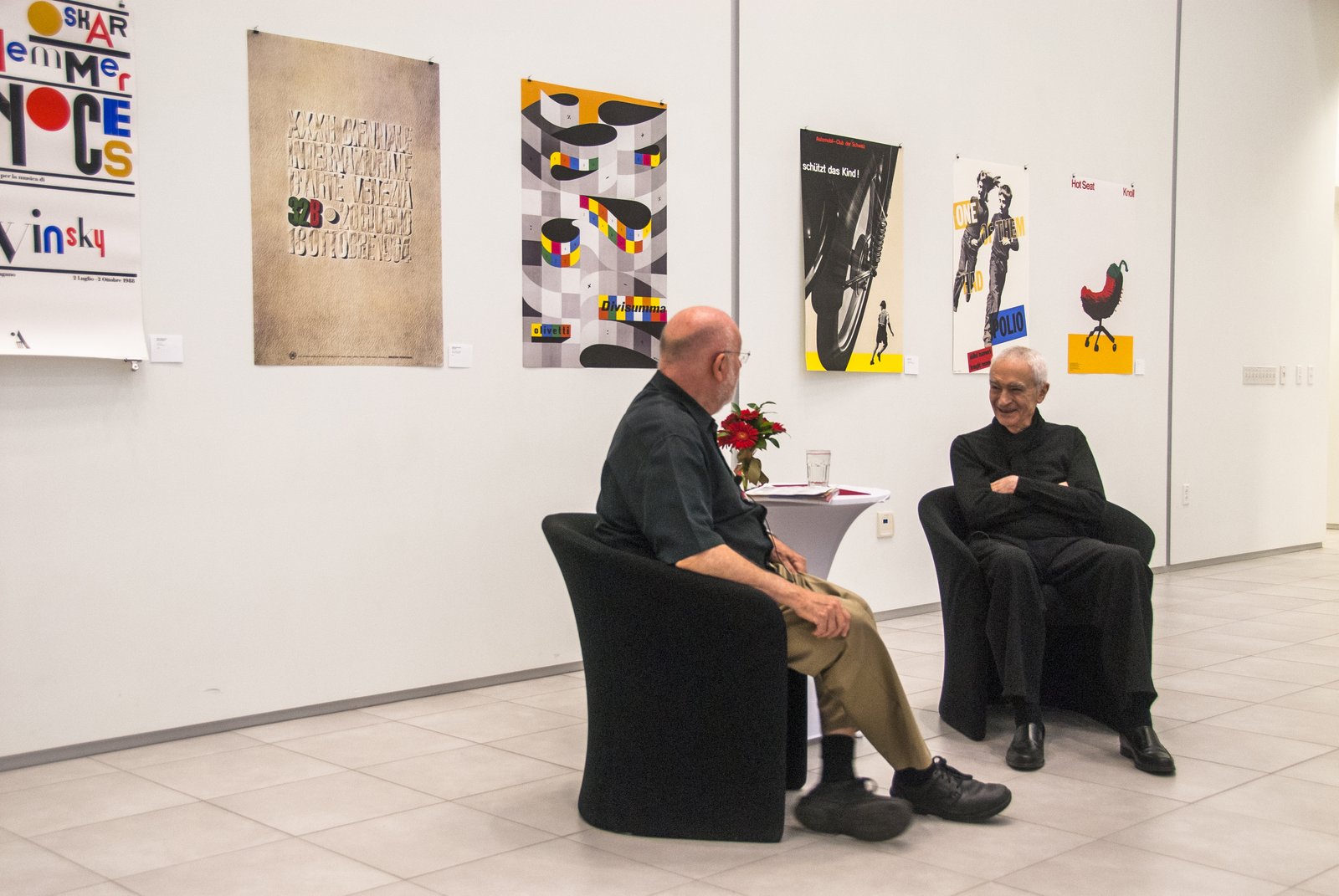 Massimo Vignelli, dressed entirely in black, seated in conversation with R. Roger Remington in the white gallery of the Vignelli Center for Design Studies, a row of his posters hung on the wall behind them