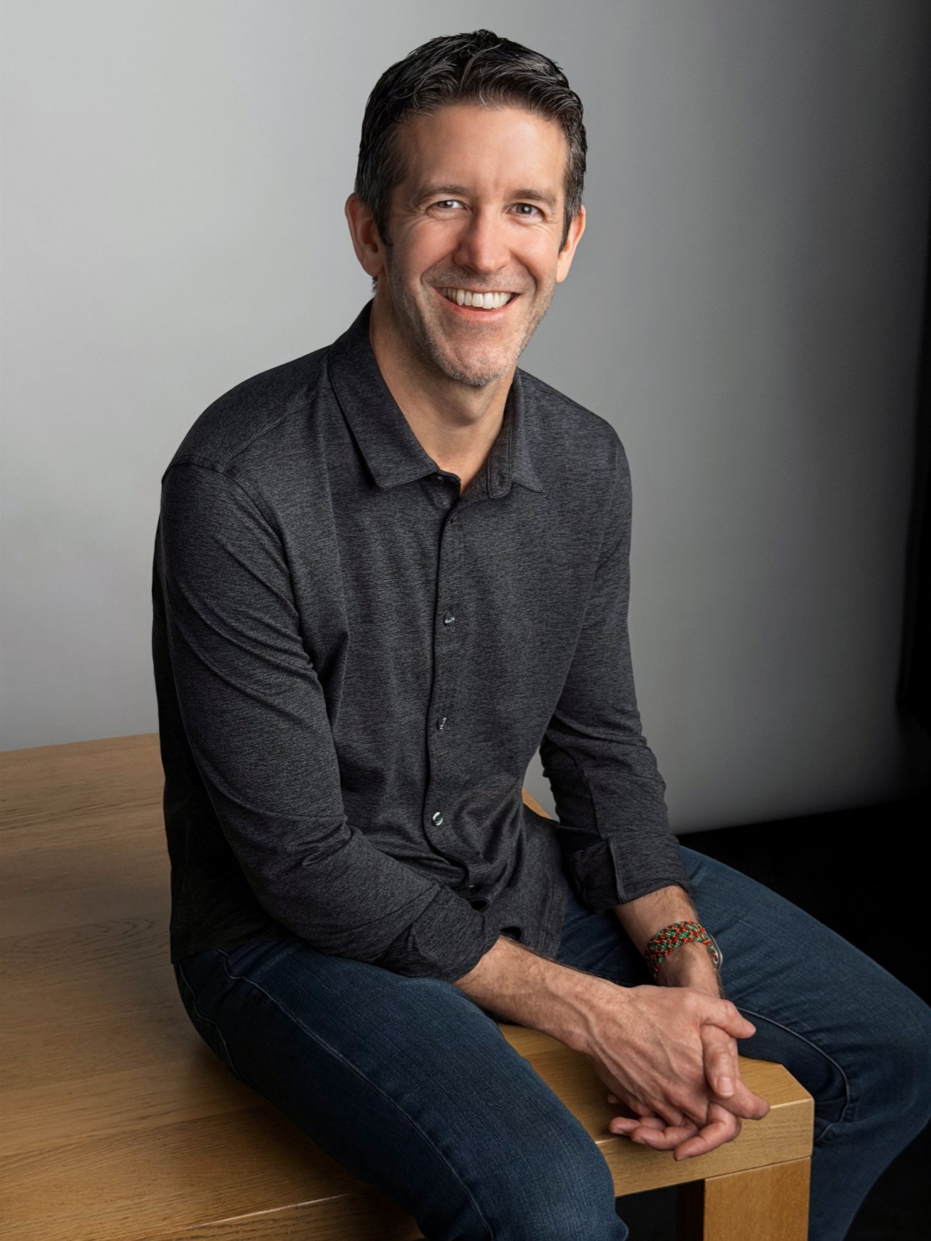 John Ternus, futur PDG d'Apple, assis sur un banc en bois, vêtu d'une chemise grise boutonnée, souriant vers la caméra.